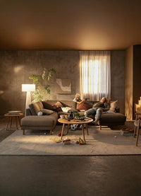 Stue med beige hjørnesofa og sofabord med snacks; et barn leger mens en voksen hviler, varm belysning, planter og trædetaljer, lidt rod på tæppet.