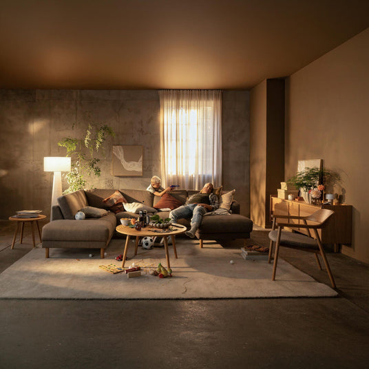 Stue med beige hjørnesofa og sofabord med snacks; et barn leger mens en voksen hviler, varm belysning, planter og trædetaljer, lidt rod på tæppet.