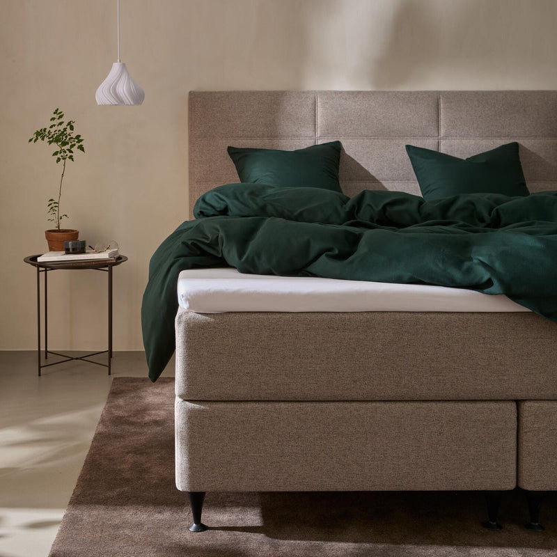 Seng med beige polstret gavl og mørkegrønt sengesæt, lille sidebord med potteplante og hvid pendellampe i et roligt soveværelse.
