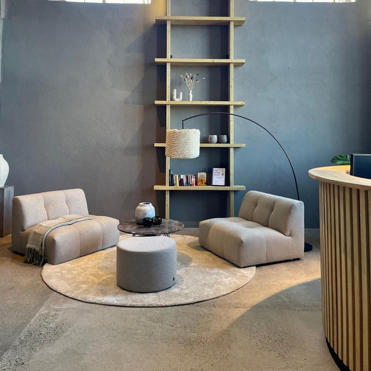 Loungehjørne med to beige loungestole, rundt tæppe, puf og sofabord foran væg med hyldesystem, buet gulvlampe og store vinduer.