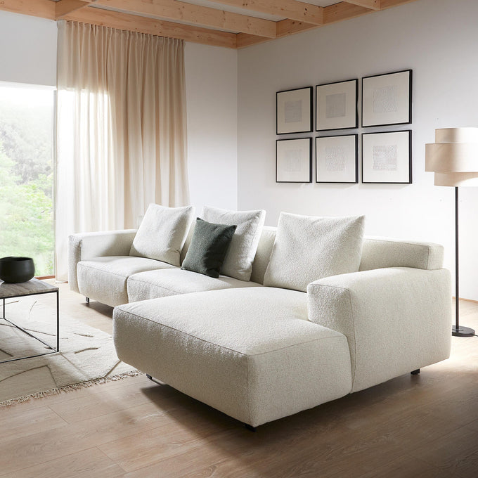 Lyst stue i skandinavisk stil med stor lys stofsofa med chaiselong, lavt sofabord, gulvlampe, vægkunst og beige gardiner med dagslys fra et stort vindue.