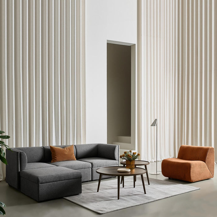 Minimalistisk stue med grå modulsofa, rustfarvet lænestol, rundt sofabord i træ med vase og blomster på lyst tæppe; høje cremefarvede gardiner og en slank gulvlampe giver et luftigt udtryk.