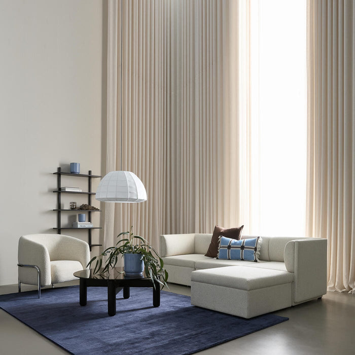 Lyst stue med beige modulsofa, blå tæppe, sort sofabord med grøn plante, pendel og høje beige gardiner.