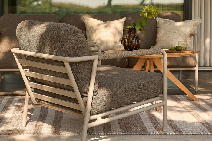 Udendørs lounge med beige metallarmlænestol, sofa og lille træbord med brun glasvase med hortensia, puder og stribet tæppe på en terrasse ved skydedøre.