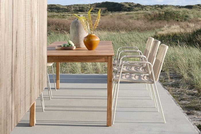 Terrasse med teakbord, vase med kviste og fire flettede stole på en betonterrasse ved klitter under blå himmel.