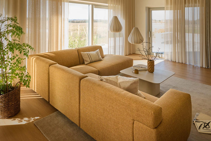 Lyst, varmt opholdsrum med sennepsgul modulsofa, lette beige gardiner og to skulpturelle pendler over et lavt lysegråt sofabord.