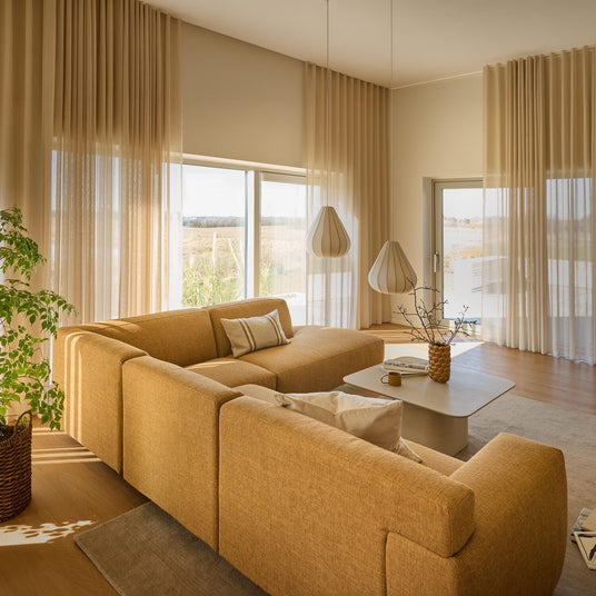 Lyst, varmt opholdsrum med sennepsgul modulsofa, lette beige gardiner og to skulpturelle pendler over et lavt lysegråt sofabord.