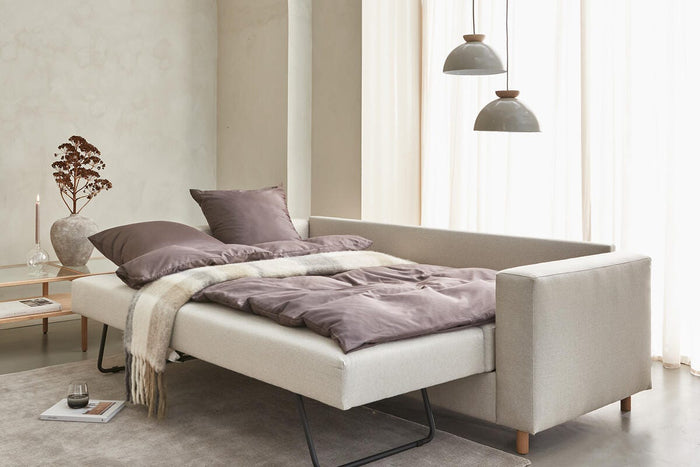 Beige sovesofa trukket ud med mauve sengetøj, to hængende pendler, lette gardiner og sidebord med vase og lys i en lys skandinavisk stue.