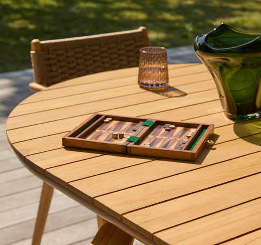 Rundt udendørs træbord med åbent backgammonspil, et glas og en grøn glasvase på en solrig terrasse.