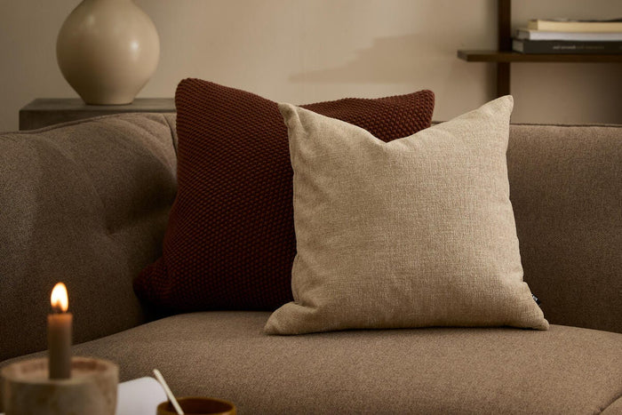 Beige pude på brun sofa med rustfarvet strikpude bagved; tændt lys, kop og bog på bordet samt vase med grønne grene og hylder i baggrunden – hyggelig stuescene.