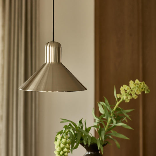 Børstet metalpendel over et bord med grønne stilke i en mørk vase i et roligt, minimalistisk rum.