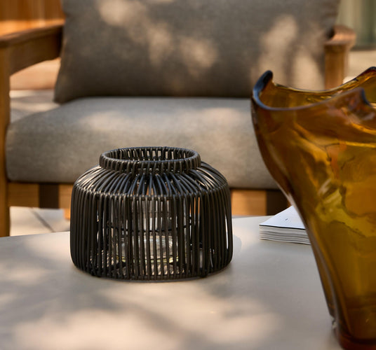 Sort lanterne i rottinglook med glasindsats på et udendørs bord, ravfarvet vase og grå sofa i baggrunden.
