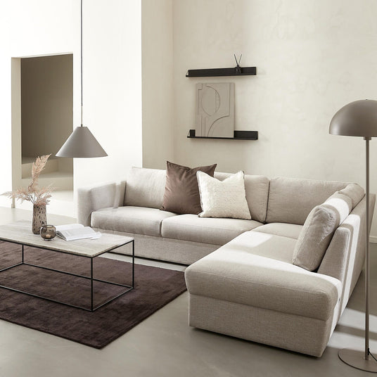 Lyst minimalistisk stue med beige hjørnesofa med chaiselong, brun tæppe, rektangulært sofabord i sten og sort metal, sort lænestol med skammel, pendel- og gulvlampe samt væghylder med abstrakt kunst.