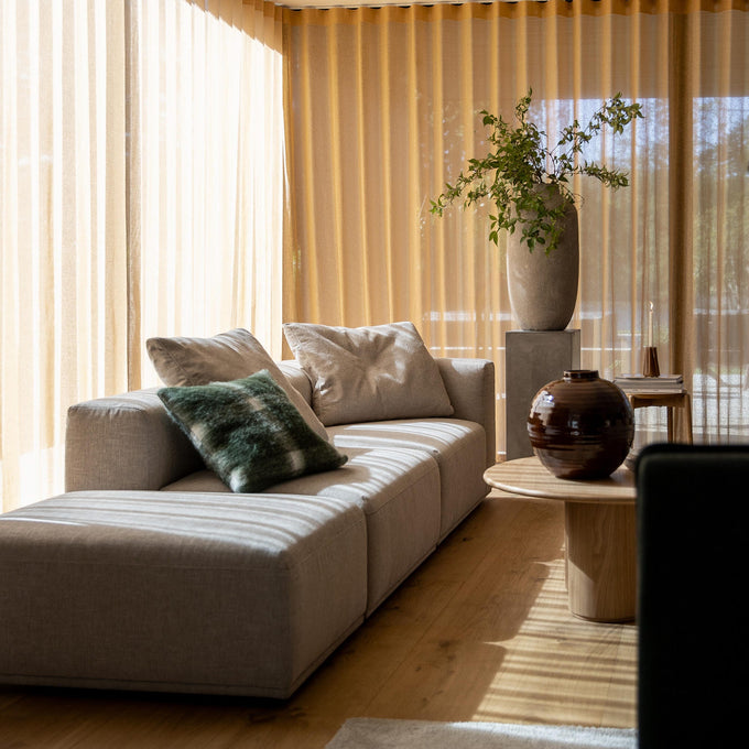 Lyst stue med beige gardiner, lysegrå modulsofa med puder, rundt sofabord i træ med brun keramisk krukke og høj vase med grønne grene på piedestal.