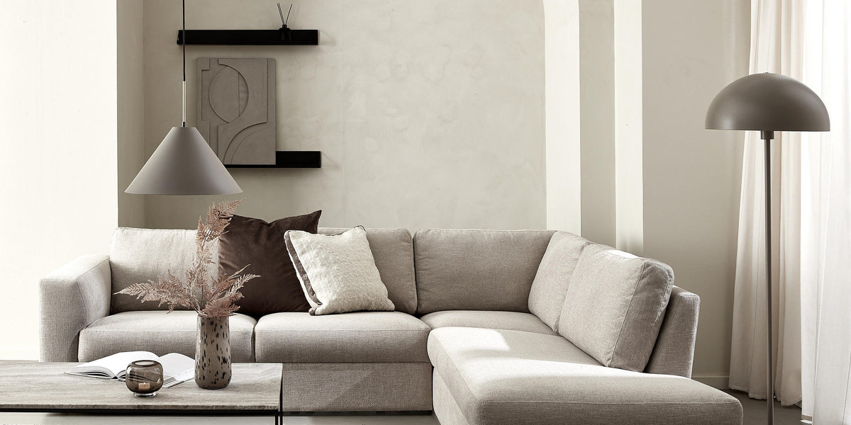 Neutralt stue med beige hjørnesofa, gulv- og pendellampe, sofabord med vase og bog, lyse gardiner og væghylder.