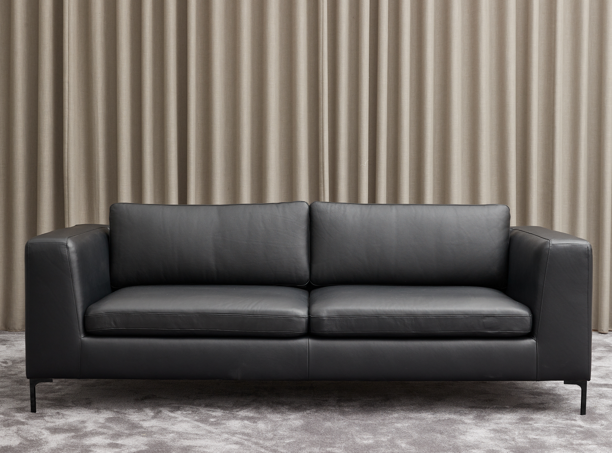 Sort lædersofa foran beige gardiner på gråt tæppe.