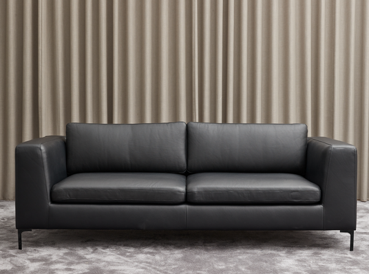 Sort lædersofa foran beige gardiner på gråt tæppe.