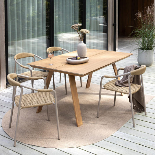 Terrasse med spisebord i træ og fire fletstole på trægulv, vase med blomster og skål på bordet, plaid over en stol og krukkeplante i baggrunden i dagslys.