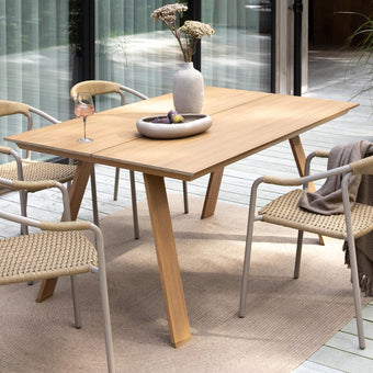 Terrasse med spisebord i træ og fire fletstole på trægulv, vase med blomster og skål på bordet, plaid over en stol og krukkeplante i baggrunden i dagslys.