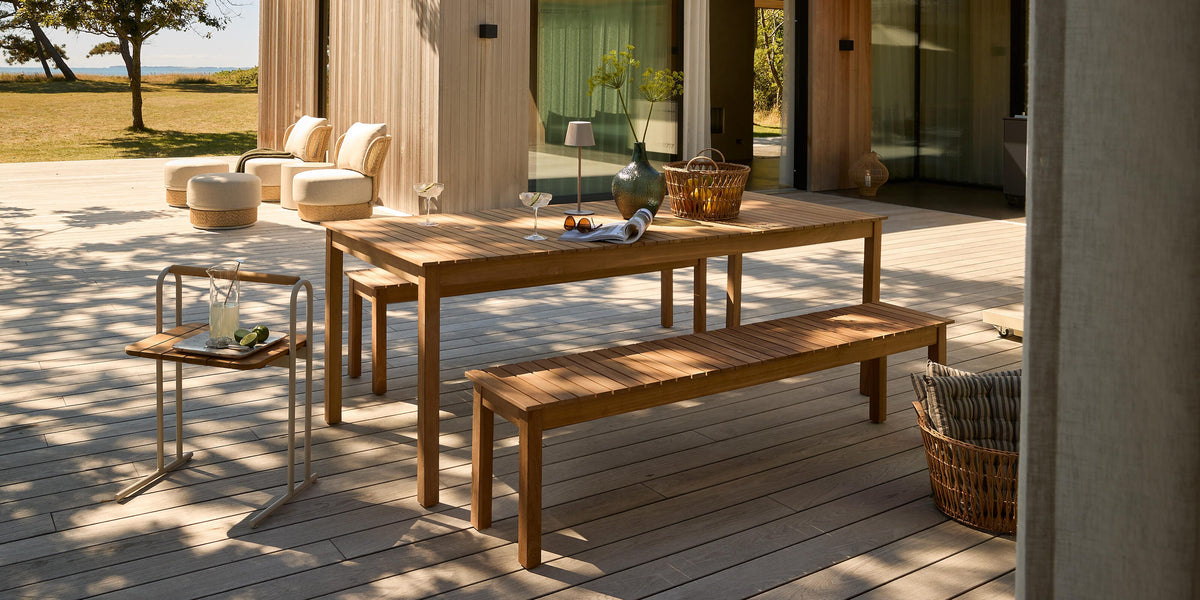 Moderne terrasse med trægulv: teakbord med bænk og lille sidebord med lemonade, loungestole og parasol foran et træbeklædt hus i solskin.