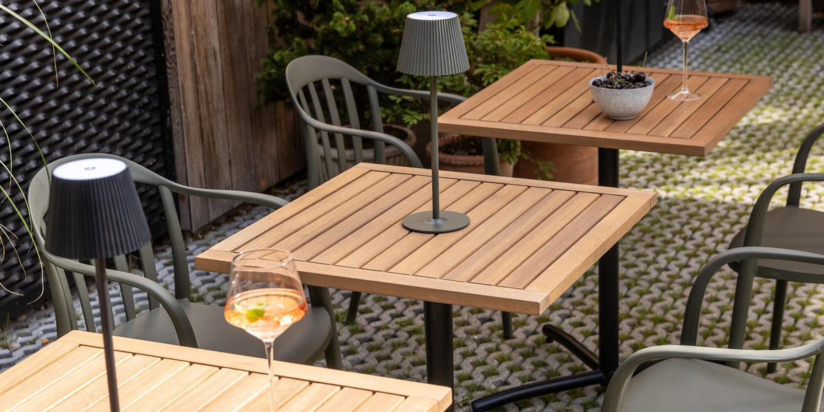 Udendørs terrasse med firkantede træborde, bærbare bordlamper og grønne stole; glas med rosé og en skål med druer i dagslys.