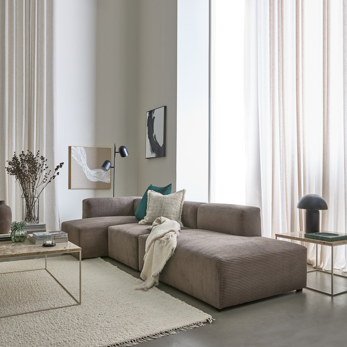 Lyst stue med beige gardiner, brun modulsofa med puder og plaid, glas- og stenborde, tæppe, vægkunst og sorte lamper.