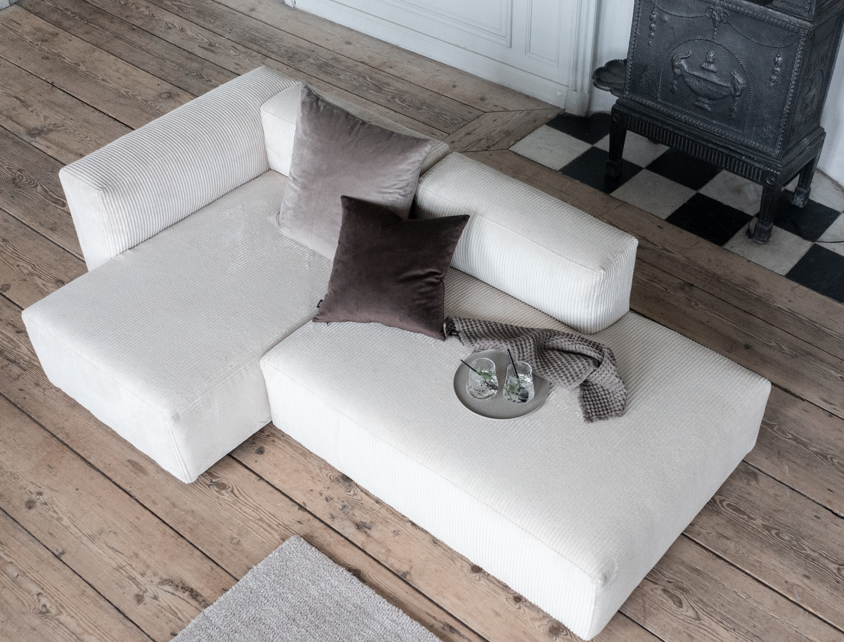 Lav modulsofa i lys beige fløjl på trægulv med to mørke puder, grå plaid og bakke med to glas; støbejernsovn i baggrunden.