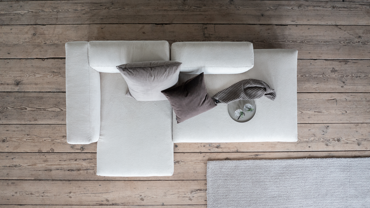 Top-down af lys beige modulsofa med to puder, strikket plaid og en bakke med glas på trægulv ved siden af tæppe i en stue.