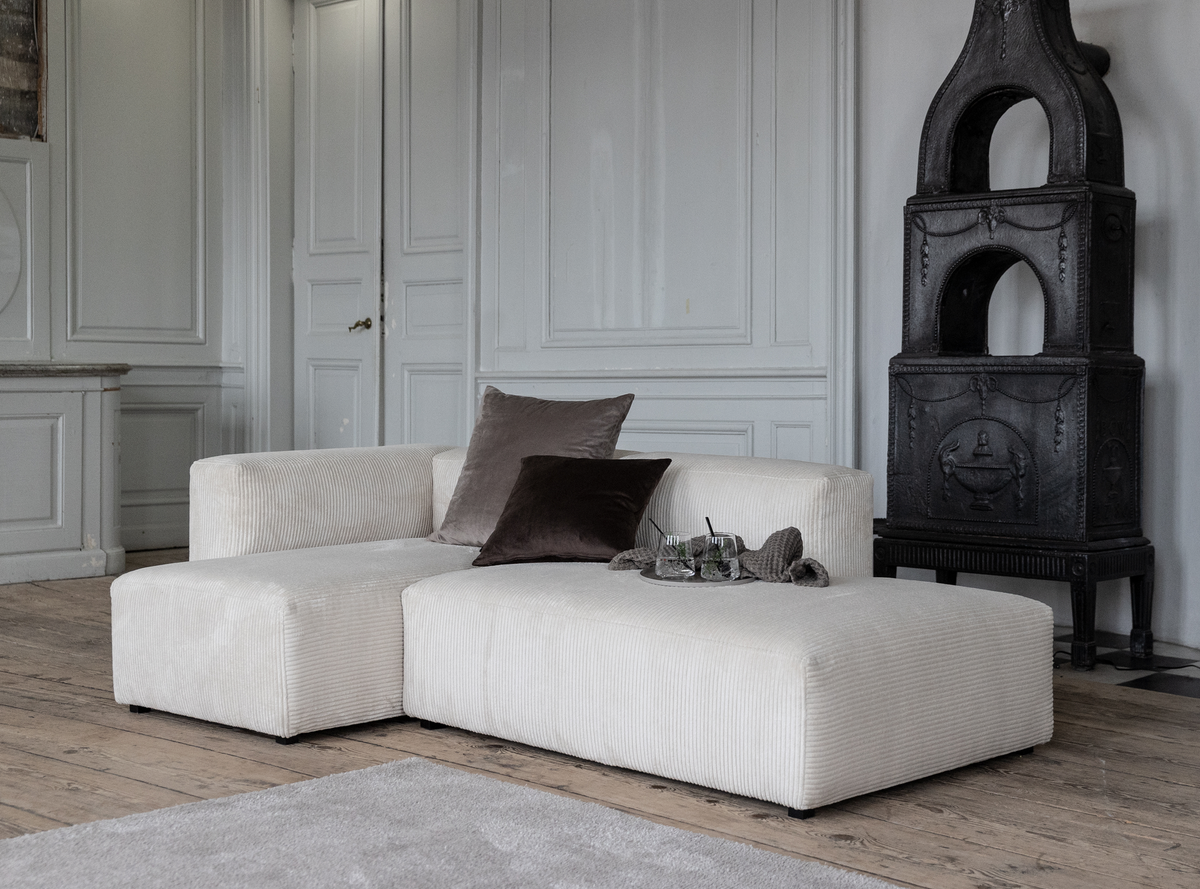 Lys L-formet modulsofa i beige fløjl med to mørke puder og en bakke med glas i en stue med grå panelvægge, plankegulv og sort støbejernsovn.