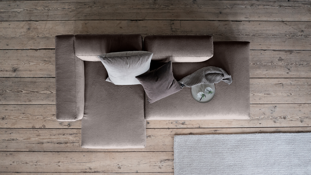 Top-down visning af en beige fløjlshjørnesofa med to puder, en strikket plaid og et fad med glas på rustikt trægulv ved siden af et lysegråt tæppe.