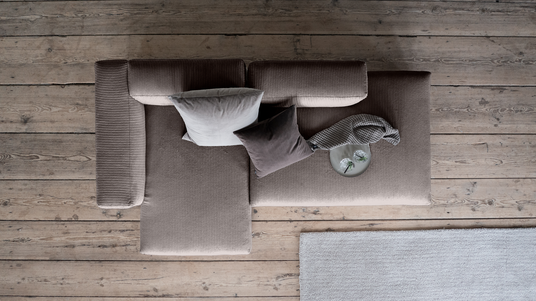 Top-down visning af en beige fløjlshjørnesofa med to puder, en strikket plaid og et fad med glas på rustikt trægulv ved siden af et lysegråt tæppe.