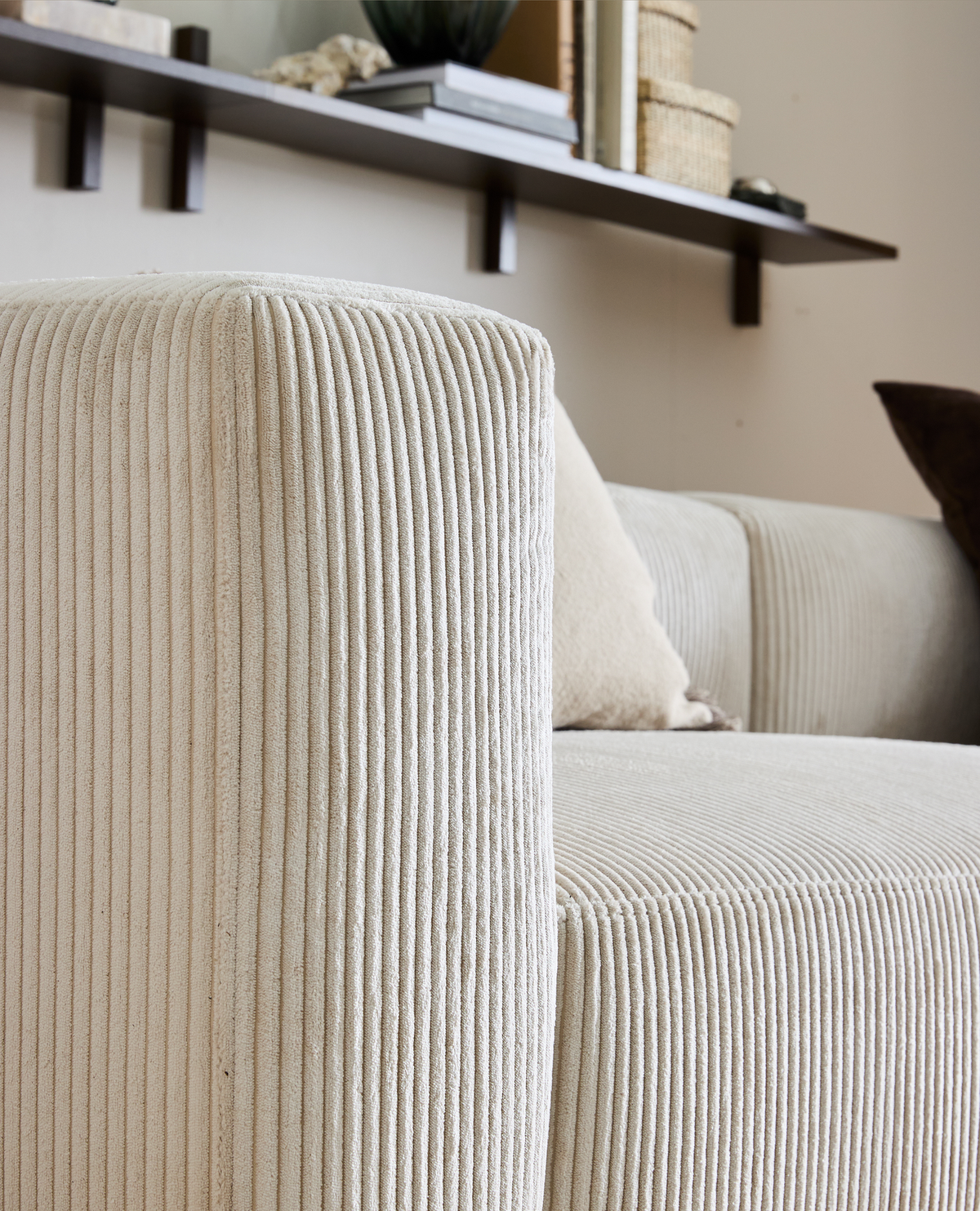 Nærbillede af beige sofa i ribbet stof med pude; væghylde med bøger, kurve og dekoration i baggrunden.
