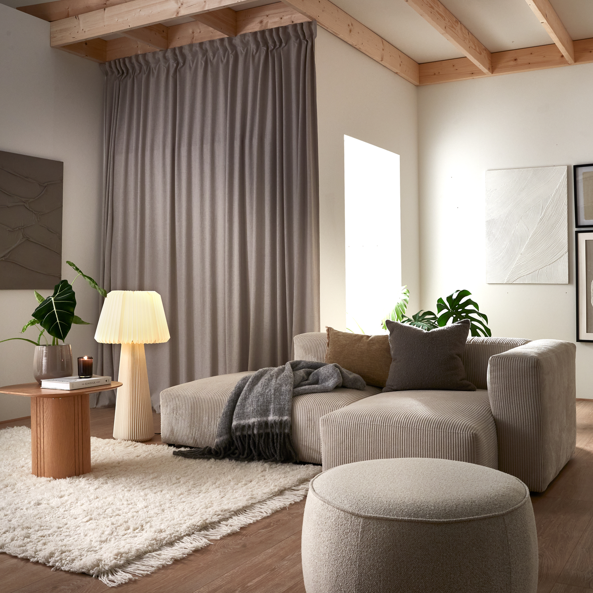 Lyst stue med beige sofa og puf, blød shaggy måtte, lille træbord med plisseret lampe, bog og plante, gardiner og minimalistisk vægkunst i neutrale toner.