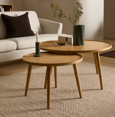 To runde indskudsborde i lyst træ på et beige uldtæppe med et tændt stearinlys, en riflede vase med grønne grene og en sofa i baggrunden.