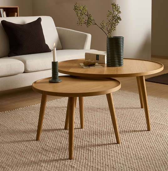 To runde indskudsborde i lyst træ på et beige uldtæppe med et tændt stearinlys, en riflede vase med grønne grene og en sofa i baggrunden.
