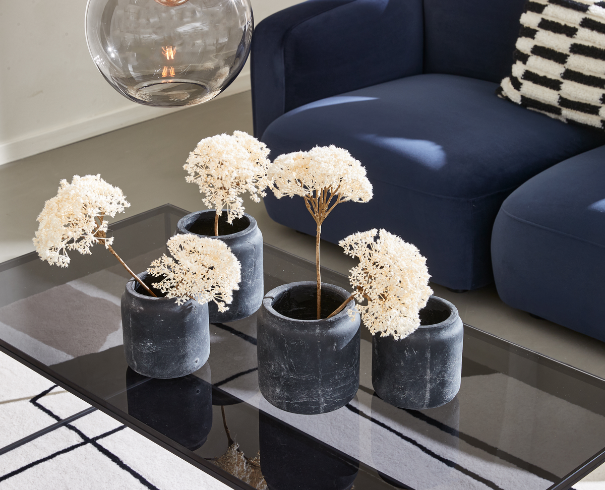 Fire sorte krukker med lyse tørrede blomster på et glasbord, blå sofa og ternet pude i en lys stue.