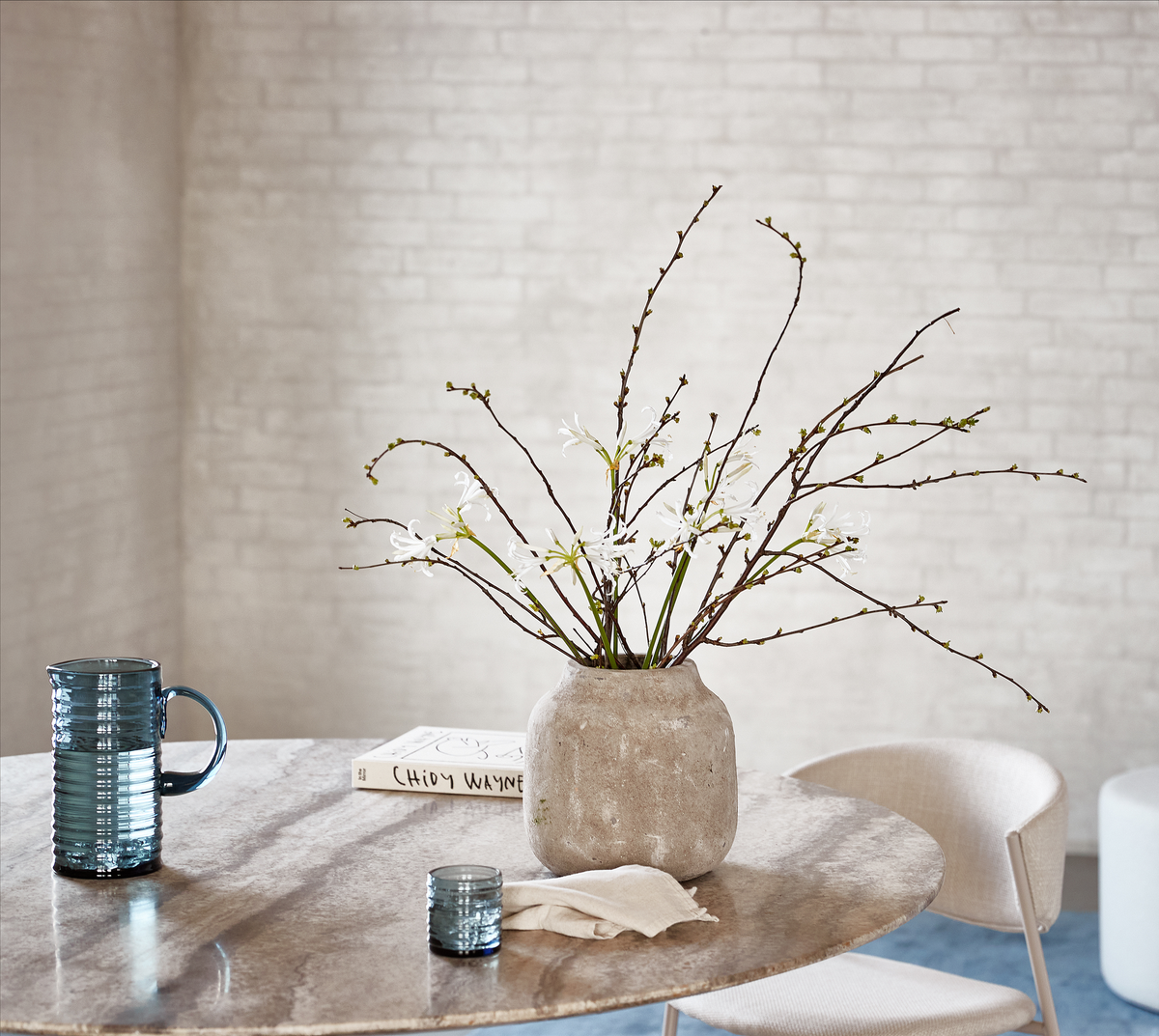 Lys og minimalistisk spiseopstilling med en beige marmorbordplade, en rustik vase med grene og hvide blomster, et blåt rillet glas og krus samt en bog med titlen ”CHOY WAYNE”.