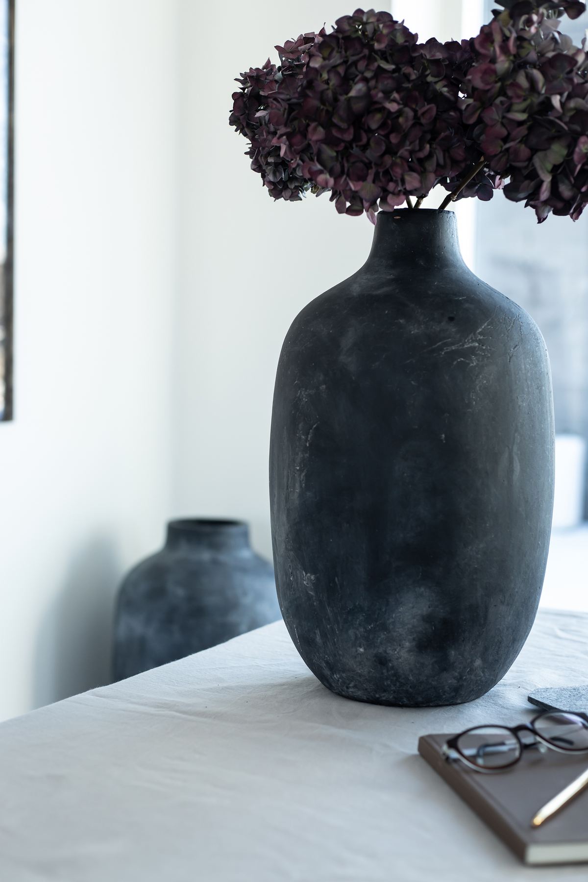 Stor mørk vase med lilla hortensia på hvid dug; notesbog med briller og pen i forgrunden i et lyst, minimalistisk indendørs miljø.