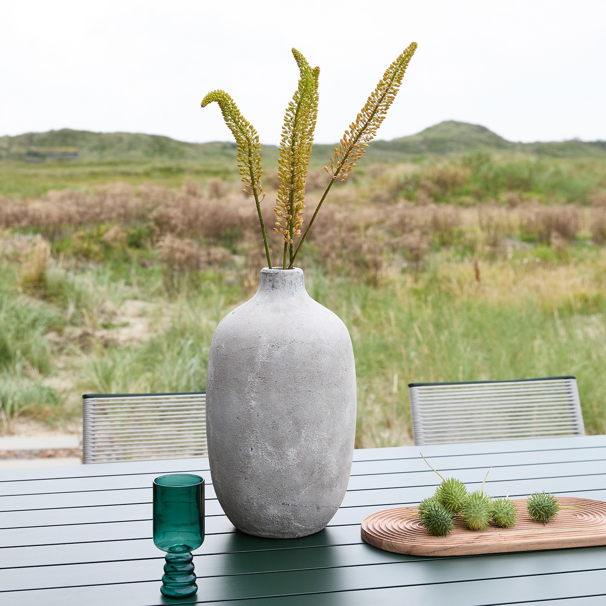 Stor grå vase med grønne stilke på et udendørs bord, med et grønt glas og et træfad med dekoration ved siden af, mod en grøn naturbaggrund.