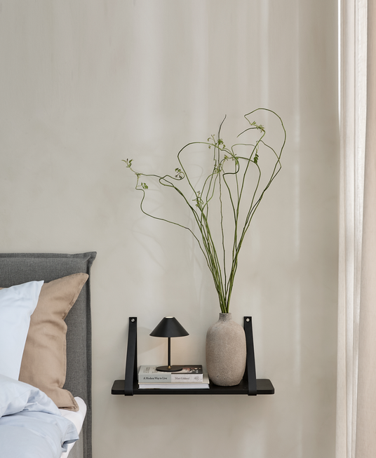 Væghylde ved sengen med en lille sort bordlampe, naturfarvet vase med lange snoede grene og en bog med titlen “A Modern Way to Live” ved siden af en grå sengegavl.