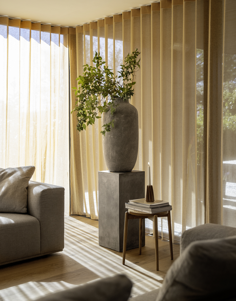 Solbeskinnet stue med gulv-til-loft beige gardiner, grå sofa, stenvase med grønne grene på sokkel og lille træbord med bøger og et lys.