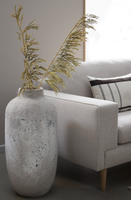 Stor betonlignende vase med tørrede strå ved siden af en lys beige sofa med pude i en lys stue.