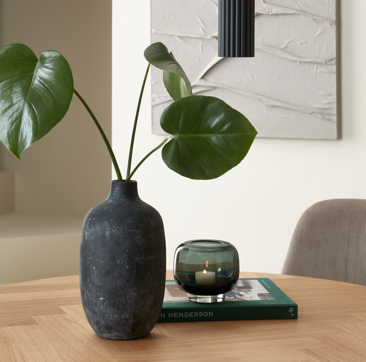 Sort vase med monstera på et træbord ved siden af en grøn glasfyrfadsstage med tændt flamme oven på en bog med “HENDERSON” på ryggen i en rolig, minimalistisk indretning.
