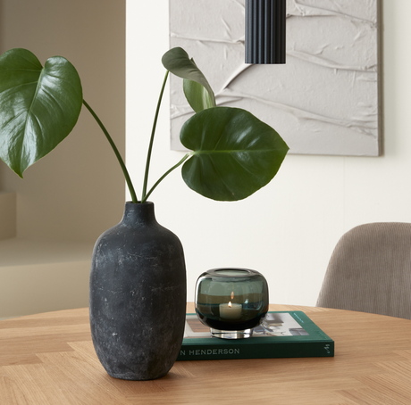 Sort vase med monstera på et træbord ved siden af en grøn glasfyrfadsstage med tændt flamme oven på en bog med “HENDERSON” på ryggen i en rolig, minimalistisk indretning.