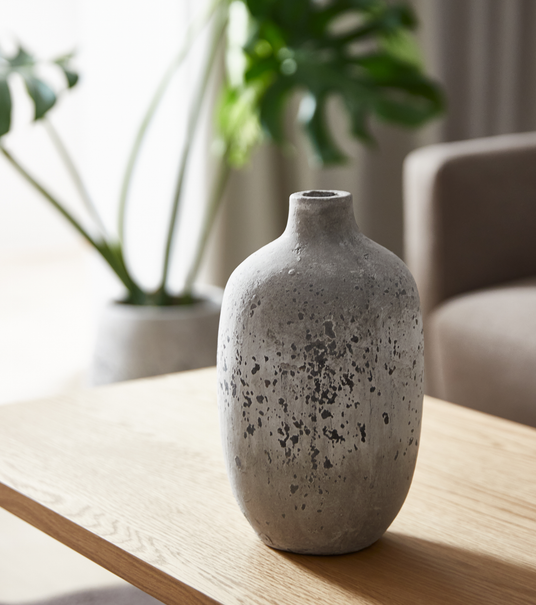 Grå, rustik keramisk vase på et lyst træbord i en stue med grønne planter sløret i baggrunden.