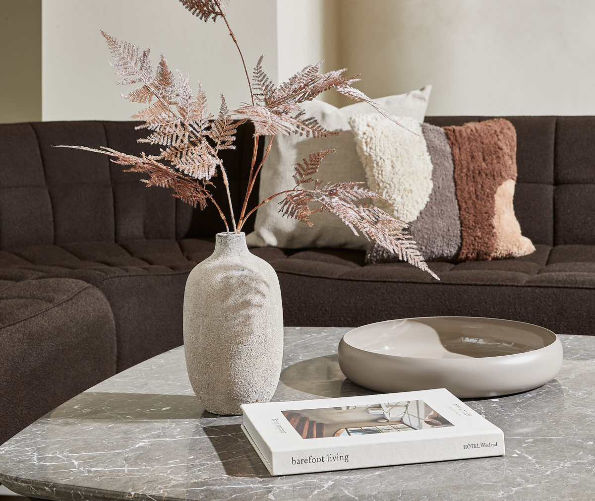 Gråt marmorbord med keramikvase med tørrede bregnegrene, lav beige skål og bogen ”barefoot living” fra HOTEL Weekend; mørkebrun sofa med teksturerede puder i baggrunden.
