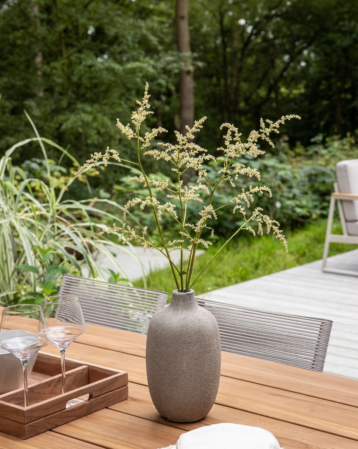 Grå keramikvase med fine græsblomster på et udendørs træbord på terrassen med grøn beplantning i baggrunden.