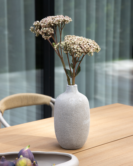 Lys grå keramikvase med hvide blomster på et udendørs træbord.