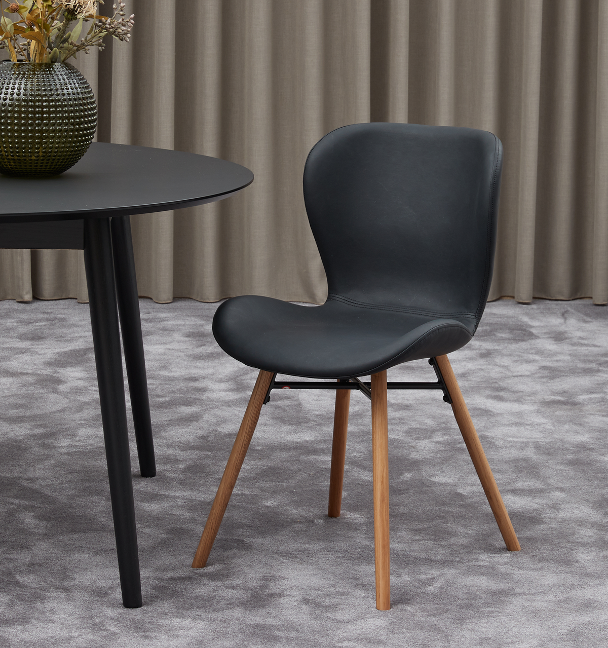 Sort spisebordsstol med træben ved siden af et rundt sort bord på et gråt tæppe, beige gardiner i baggrunden og en grøn vase med tørrede grene på bordet.
