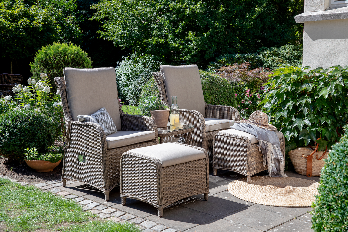 Havescene med to loungestole i flet med beige hynder og matchende skamler, lille sidebord med lemonade, stråhat og plaid på en solbeskinnet brolagt terrasse omgivet af grønne buske.